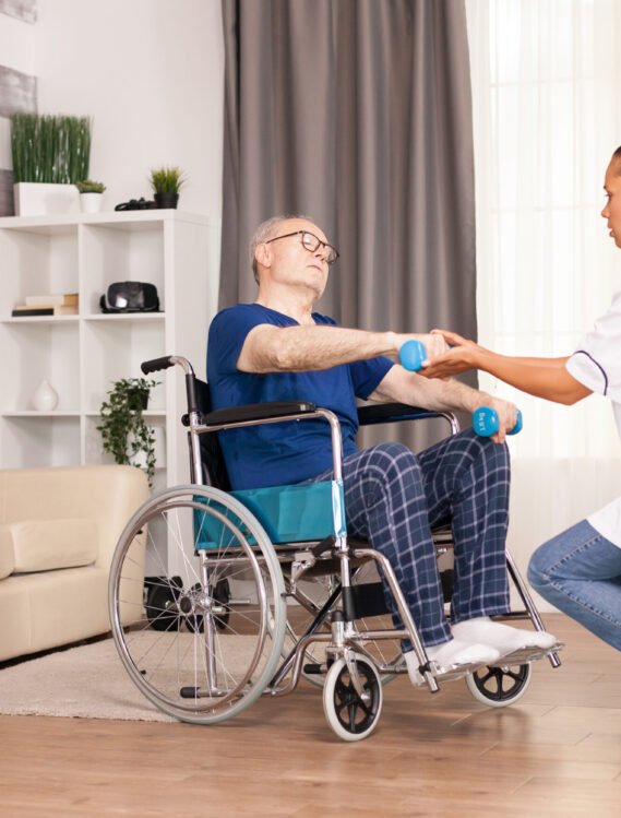Aged person suffering arms pain sitting on wheelchair and training with dumbbells. Disabled handicapped old person with social worker in recovery support therapy physiotherapy healthcare system nursing retirement home
